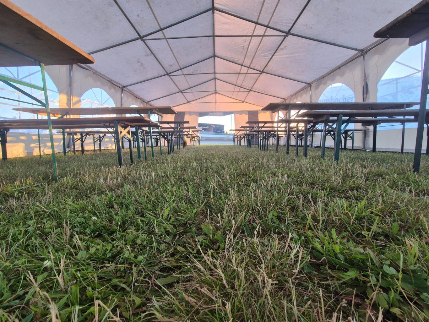 Event Großes Zelt mit mehreren Banktischen auf einem sandigen Platz, umgeben von Bäumen.